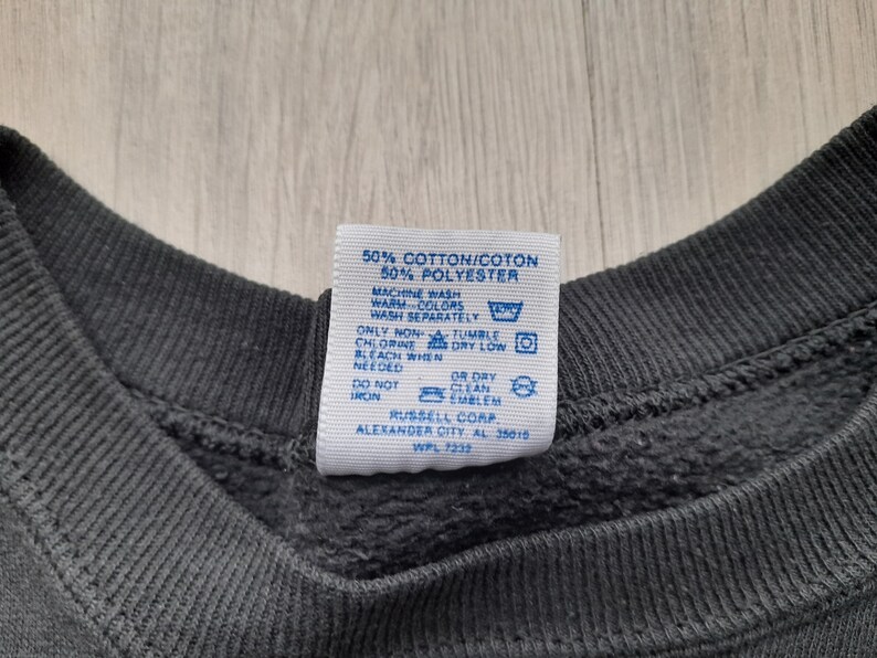 May include: A blue care label with white text detailing the fabric composition and care instructions for a garment. The label reads: "50% Cotton/Coton 50% Polyester Machine Wash Warm Colors Wash Separately Chlorine Bleach When Needed Do Not Iron Or Dry Clean Emblem Russell Corp Alexander City AL 35015 WPL 7232"