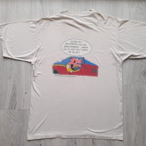 Puede incluir: Camiseta de color crema con una ilustración de dibujos animados de un personaje en un coche rojo y azul. Una burbuja de diálogo sobre el coche contiene texto en alemán. La camiseta está sobre una superficie de madera.