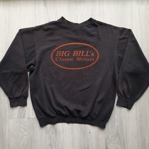 以下が含まれることがあります： オレンジ色の楕円の中に「BIG BILL'S Classic Motors」の文字が入った濃いグレーのスウェットシャツ。長袖のクルーネックシャツは柔らかい素材でできています。ヴィンテージスタイルのスウェットシャツは、明るい木材の表面に平らに置かれています。