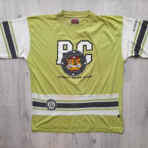 May include: Green t-shirt with a white and black striped design. The shirt features a graphic of a cartoon dog wearing a hat and the text "RC Street Wear Club".