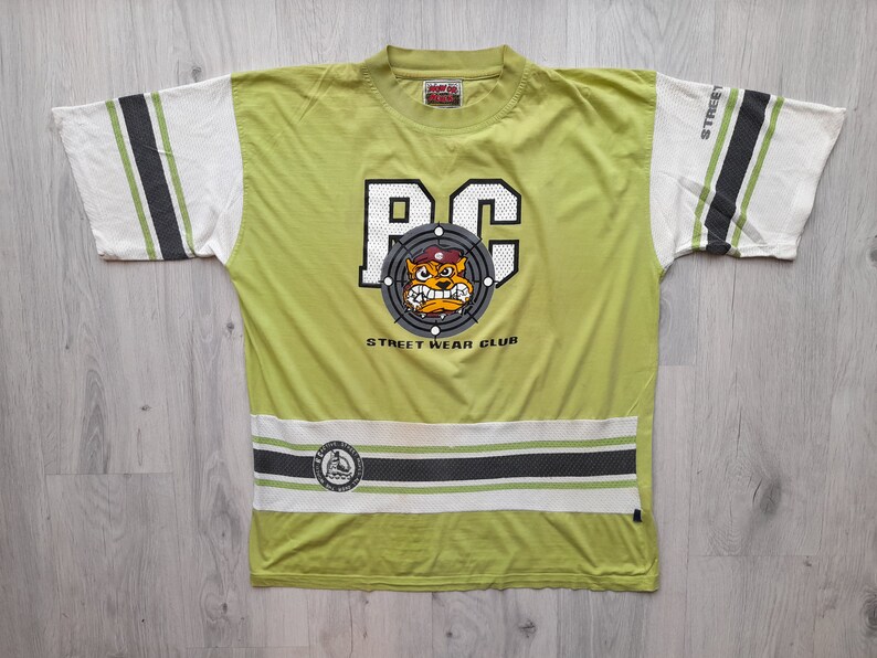 May include: Green t-shirt with a white and black striped design. The shirt features a graphic of a cartoon dog wearing a hat and the text "RC Street Wear Club".