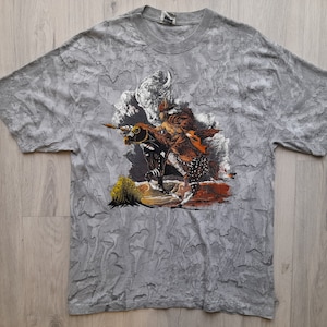 Peut inclure: T-shirt gris avec un imprimé graphique d'un guerrier amérindien à cheval. Le t-shirt a un aspect texturé et vieilli.