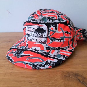 Puede incluir: Una gorra de béisbol con estampado rojo, negro y blanco con un parche que dice "Sanjaya Collection Bali Tanah Lot".