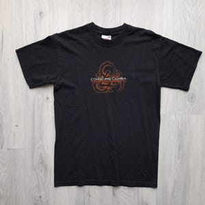 Può includere: T-shirt nero con un disegno circolare e la scritta "COHEED AND CAMBRIA" in bianco e arancione. La maglietta a maniche corte è realizzata in un materiale morbido. La maglietta è stesa su una superficie di legno chiaro.