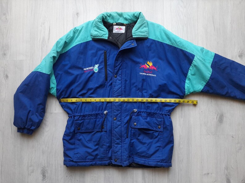 Puede incluir: Chaqueta vintage azul y turquesa con el texto "PETRONAS" y un logotipo. La chaqueta tiene cierre de cremallera, dos bolsillos delanteros y una cinta m&eacute;trica amarilla en la cintura. La chaqueta es talla L.