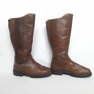 Puede incluir: Un par de botas de montar de cuero marrón. Las botas son hasta la rodilla con punta redonda y tacón bajo. Tienen cremallera en la parte trasera y un panel lateral. Las botas están hechas de cuero liso y pulido.