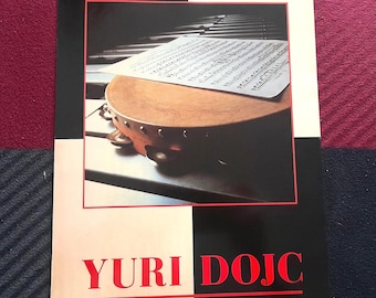 Op de afbeelding: Een poster met een close-up van een tamboerijn op een pianoklavier, met bladmuziek erop. De poster heeft een rode en zwarte rand, met de naam "YURI DOJC" in rode letters onderaan.