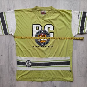 May include: A green t-shirt with a white and black striped design on the sleeves and bottom. The shirt features a large graphic of a cartoon bulldog wearing a hat and a target with the letters "BC" in the center. The text "STREET WEAR CLUB" is printed below the graphic.