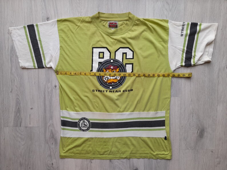May include: A green t-shirt with a white and black striped design on the sleeves and bottom. The shirt features a large graphic of a cartoon bulldog wearing a hat and a target with the letters "BC" in the center. The text "STREET WEAR CLUB" is printed below the graphic.