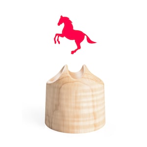 Puede incluir: Un soporte de madera de color claro con una base cilíndrica lisa y una parte superior que presenta tres hendiduras curvas distintas. Sobre el objeto de madera flota una silueta sólida de color rosa brillante de un caballo encabritado.