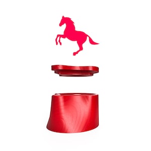 May include: A red bottle stopper with a horse silhouette design. The stopper is made of three parts: a base, a middle piece, and a horse silhouette. The base is a cylindrical shape. The horse silhouette is above the stopper.