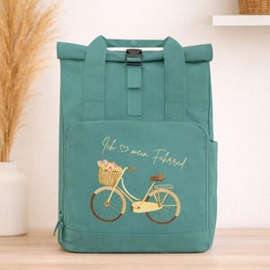 Könnte beinhalten: Türkisfarbener Rucksack mit Rollverschluss und einer Vordertasche. Auf der Vorderseite des Rucksacks befindet sich eine Fahrradillustration mit einem Blumenkorb und dem Text "Ich mein Fahrrad".