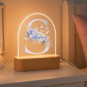 May include: An arched acrylic night light featuring a large white letter S decorated with blue floral illustrations and the name Susanne in cursive script. The clear panel is set into a rectangular light-colored wooden base that emits a warm glow.