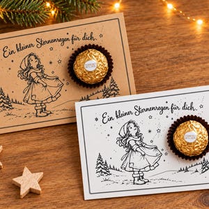 Può includere: Due cartoline rettangolari con un disegno a tratto nero di un bambino in abiti invernali sotto le stelle, con il testo tedesco "Ein kleiner Sternenregen für dich." Ogni cartolina contiene un cioccolatino Ferrero Rocher dorato.