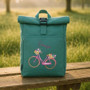 Pode incluir: Uma mochila verde-azulado com fecho enrolável e fivela preta. A frente apresenta um desenho de bicicleta rosa com flores e o nome "Vanessa" em itálico. A mochila está em um banco de madeira.