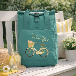 Puede incluir: Mochila verde azulado con una ilustración de bicicleta y el texto "Fahrradglück" en el bolsillo delantero. La bolsa tiene un asa superior y un cierre de hebilla. Un vaso de limonada, un sombrero de paja y un libro están en un banco.