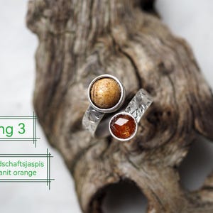 Könnte beinhalten: Handgefertigter Silberring mit zwei Edelsteinfassungen. Eine Fassung hält einen runden, beigefarbenen Stein, die andere einen facettierten, orangeroten Stein. Der Ring wird auf einem Stück Treibholz präsentiert. Der Text auf dem Bild lautet "Ring 3" und "Landschaftsjaspsis Kyanit orange".