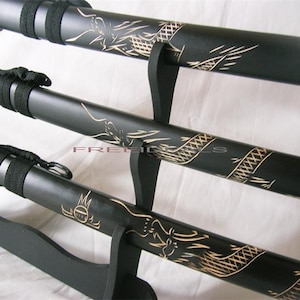 May include: Three black katana swords with gold dragon designs on the scabbards are displayed on a black stand.