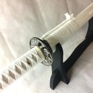 May include: A Japanese katana sword with a white handle and blade, displayed on a black stand. The handle is wrapped in white fabric with a diamond pattern. The sword's guard is silver with a detailed design.