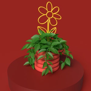 May include: A red planter with a green plant and a yellow flower stake. The planter has a ribbed design and sits on a red pedestal, against a red backdrop. The plant's green leaves cascade over the sides of the planter, creating a decorative display.