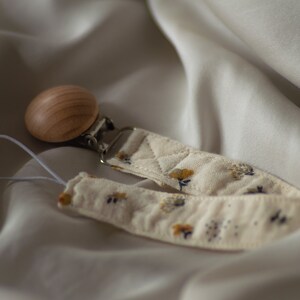 Peut inclure: Une sucette en bois avec une sangle en tissu à motif floral et un clip en métal. La sangle est de couleur crème avec de petites fleurs jaunes et bleues.