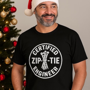 Puede incluir: Un hombre con una camiseta negra con un gráfico circular blanco que dice "CERTIFIED ZIP-TIE ENGINEER". También lleva un gorro de Papá Noel rojo. Un árbol de Navidad con adornos rojos y dorados está en el fondo.