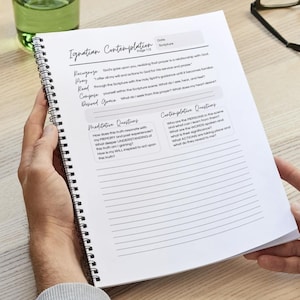 May include: A spiral-bound journal with the title "Ignatian Contemplation" is open, revealing lined pages and text. The journal is held by hands, with a glass of water and eyeglasses in the background. The journal includes prompts for reflection.