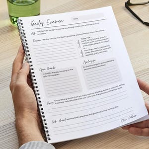 Peut inclure: Un carnet à spirales intitulé "Daily Examen" avec des invites à la réflexion. Les sections comprennent demander, revoir, remercier, s'excuser, prier et regarder vers l'avenir. Le carnet est tenu dans les mains de quelqu'un. Le texte est manuscrit.