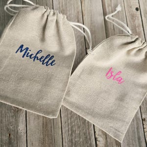 May include: Two beige drawstring bags with white cords. One bag has the name "Michelle" embroidered in blue, and the other has "Isla" in pink. The bags are made of a textured fabric.