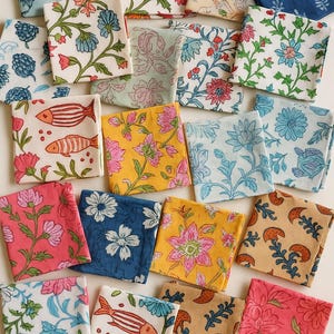May include: A collection of folded, square fabric napkins with various floral and botanical patterns. The napkins feature a range of colors, including blue, pink, yellow, and white. Some designs include fish motifs. The napkins are likely for home decor.