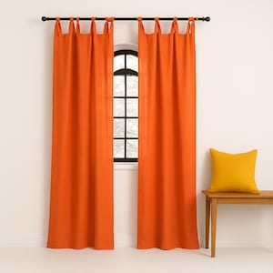 Puede incluir: Cortinas naranjas brillantes con presillas colgadas de una barra oscura, enmarcando una ventana con una rejilla negra. Un cojín amarillo descansa sobre un banco de madera. Las cortinas son de color liso y parecen estar hechas de tela texturizada.