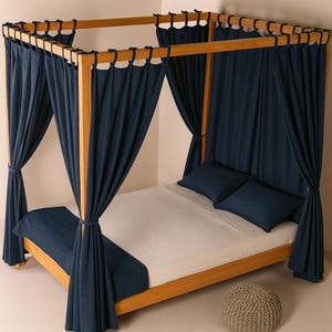 May include: A wooden four-poster bed with navy blue curtains and matching pillows. The bed frame is made of light-coloured wood. A grey knitted pouf sits at the foot of the bed. The bed is made up with a white sheet and a navy blue blanket.