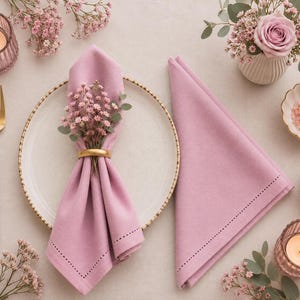 May include: A table setting featuring two mauve cloth napkins with hemstitch detailing, a white plate with a gold rim, a gold napkin ring holding baby's breath, a gold and mauve fork, two pink glass candle holders, and small bowls of spices.