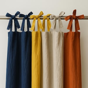 May include: Five linen curtain panels in navy blue, yellow, beige, and rust orange hang from a silver rod. Each panel is tied with a matching fabric bow at the top. The curtains are made of a soft, textured fabric.