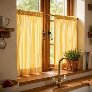 Könnte beinhalten: Ein Küchenfenster mit gelb-weiß gestreiften Vorhängen. Vor dem Fenster befindet sich ein Wasserhahn aus Messing. Eine kleine Topfpflanze und Bücher stehen auf der Holzfensterbank. Ein Holzregal hält Tassen.