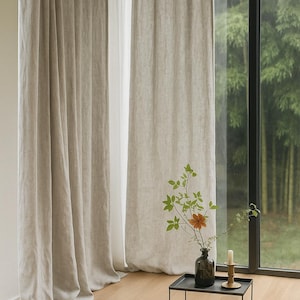 May include: Neutral linen curtains hang beside a large window with a view of a green bamboo forest. A small black metal side table holds a vase of flowers, a candle, and a round floor cushion. A decorative log lies on the wooden floor.