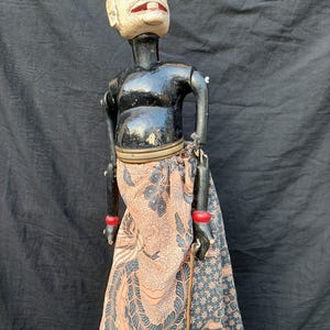 Peut inclure: Une marionnette traditionnelle indonésienne Wayang Golek. La marionnette a un visage blanc avec des lèvres rouges, un corps noir et une jupe à motifs dans les tons pêche, bleu et noir. Les bras de la marionnette sont articulés.