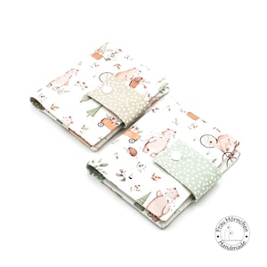 Könnte beinhalten: Zwei weiße Stoffkartenhalter mit einem Bären- und Waldmotiv. Der kleinere Halter hat eine grün-weiß gepunktete Klappe mit einem weißen Knopf. Der größere Halter hat eine beige-weiß gepunktete Klappe mit einem weißen Knopf. Der Text "Frau Hörnchen Handmade" ist auf einem weißen Kreis gedruckt.