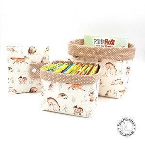 Könnte beinhalten: Drei Stoffaufbewahrungskörbe mit einem Waldtierdruck. Der größte Korb ist mit Büchern gefüllt und hat den Text "Ritter Rost und der Drache" auf dem obersten Buch. Die Körbe sind beige mit braunen Punkten.