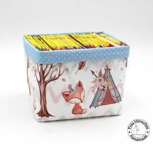 Könnte beinhalten: Eine blaue und weiße Stoffaufbewahrungsbox mit einem Waldtierdruck. Die Box ist mit bunten Büchern gefüllt und zeigt einen Fuchs, einen Baum und ein Tipi. Die Box hat einen blauen Tupfenrand und die Worte "Frau Hörnchen Handmade" sind auf dem Boden aufgedruckt.