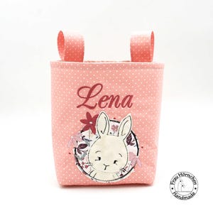 Könnte beinhalten: Rosa Stoffaufbewahrungskorb mit weißen Punkten, mit einem personalisierten bestickten Namen "Lena" und einem niedlichen Hasen-Applikationen mit Blumendetails.