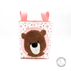 Könnte beinhalten: Ein rosa Stoffaufbewahrungskorb mit einem braunen Bärenkopf-Applikationen. Der Korb hat rosa Griffe und einen weißen Hintergrund mit rosa Blümchenmuster. Der Text "Frau Hörnchen Handmade" befindet sich am Boden des Korbes.