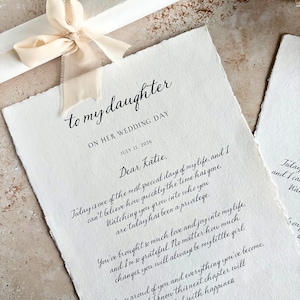 May include: Off-white wedding letter with calligraphy text, addressed "to my daughter" with the date July 11, 2026. A cream-colored ribbon is tied around a rolled-up paper. The letter's text is a heartfelt message.