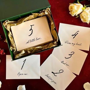 Op de afbeelding: Een groene doos met goudkleurig vloeipapier bevat kaarten met handgeschreven berichten. Vijf witte kaarten zijn zichtbaar, elk met een nummer en een korte boodschap in zwart schrift: "Open me", "Just for you", "Read me", "From my heart" en "A little love."