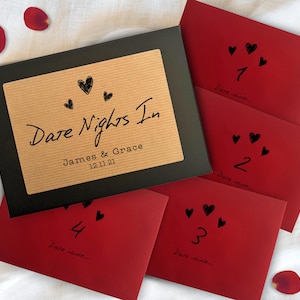 Può includere: Una scatola nera con un'etichetta marrone che recita "Dare Nights In", i nomi "James & Grace" e la data "12.11.21". Sono visibili anche quattro buste rosse con cuori neri e numeri da 1 a 4. Petali di rosa sparsi.