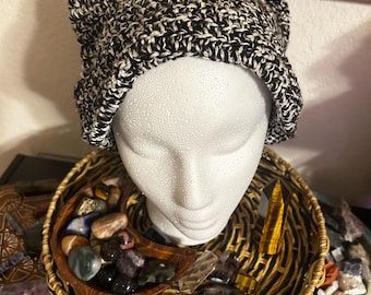 May include: Black and white crocheted beanie with cat ears. The beanie is on a white mannequin head. The mannequin head is on a wicker basket filled with crystals.