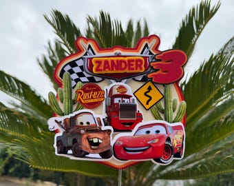 Puede incluir: Un adorno para pastel de Cars de color rojo, amarillo y azul con el nombre "Zander" y el número "3". El adorno presenta un coche de carreras rojo, una grúa oxidada y un camión rojo con la palabra "Rust-eze" en él.