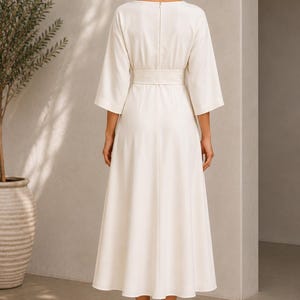 May include: A cream-colored midi dress featuring three-quarter length sleeves, a wide waistband, and a back zipper closure. The garment has a structured bodice and a flowing skirt that falls below the knees, paired with tan high-heeled sandals.