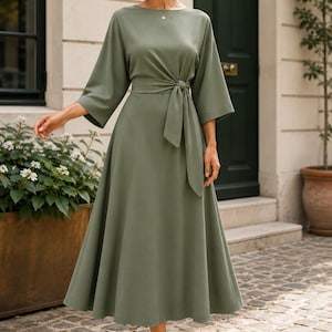 May include: A sage green midi dress featuring a round neckline, three-quarter length sleeves, and a waist tie detail. The dress has a fitted bodice that transitions into a flared skirt, reaching below the knees, paired with neutral-toned heels.