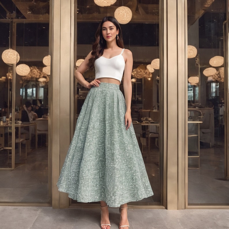 May include: A woman wearing a white crop top and a green floral midi skirt. She is standing in front of a glass door with gold trim.